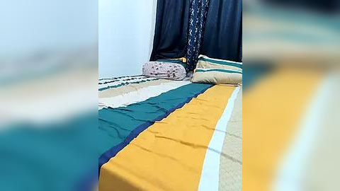 Media: Video of a bed with a colorful striped bedspread featuring yellow, teal, and white stripes, against a dark blue curtain backdrop, with a decorative pillow in the foreground.