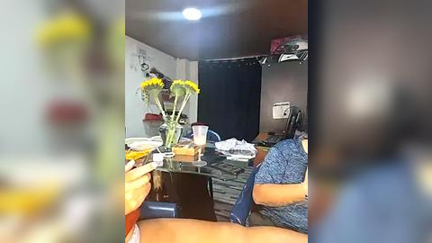 Media: A video shows a messy dining table with a vase of sunflowers, a blue shirt, and a blurry figure in the foreground. The background features a dimly lit room with a black curtain and a cluttered shelf.