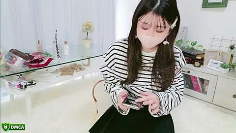 Media: Video of a woman with long black hair, wearing a white face mask, black-and-white striped sweater, and black skirt, examining a small device in a brightly lit room.