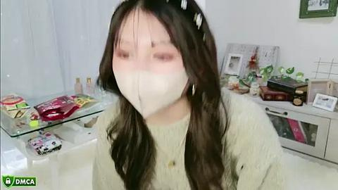 Media: Video of an Asian woman with long black hair, wearing a white mask, a beige sweater, and sitting in a bright, cluttered kitchen.