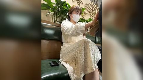 Media: Video of a woman with fair skin, brown hair, wearing a white mask, cream lace dress, and sitting on a green leather couch.