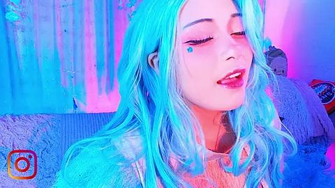 Media: Video of a young woman with long, vibrant blue hair, wearing a white sweater, and makeup with a blue heart on her cheek, set against a colorful, abstract background.