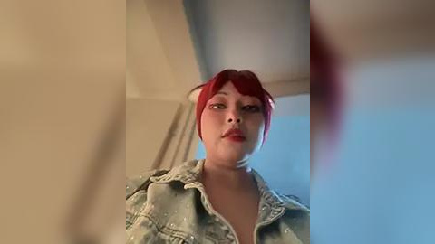 Media: Video of a young woman with red hair and fair skin wearing a denim jacket, standing in a dimly lit room with beige walls and a window.