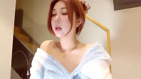 Media: Video of a young Asian woman with fair skin, auburn hair, wearing a light blue off-shoulder sweater, standing on a staircase with a beige wall background.