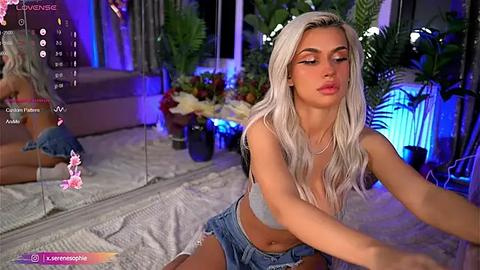 Media: Video of a blonde, light-skinned woman in a gray crop top, denim shorts, and a floral headband, sitting on a bed with lush green plants and vibrant flowers in the background.