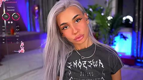 Media: Video of a young, light-skinned woman with long, platinum blonde hair and sharp, black eyebrows. She wears a black t-shirt with white graffiti-like text and a silver necklace. Background features a modern, dimly-lit room with green plants and blue lighting.