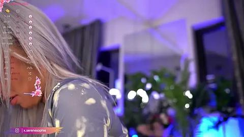 Media: Video of a woman with long, silver hair, wearing a shiny, metallic silver outfit, standing indoors with a blurred background featuring green plants and purple lighting.