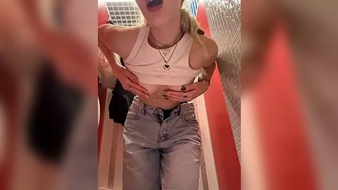 Media: Video of a slender, fair-skinned woman with long blonde hair, wearing a white crop top, blue jeans, and a heart necklace, in a red-tiled bathroom, mouth open, possibly screaming.