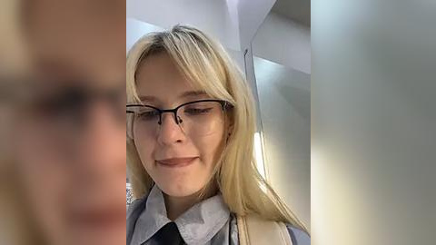 Media: Video of a blonde woman with glasses, smiling, wearing a light blue shirt, standing in an airport terminal with modern architecture.