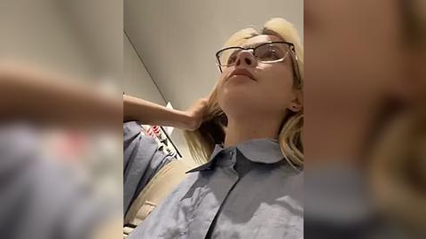 Media: Video of a blonde woman with glasses, wearing a blue shirt, leaning back with hands on her head, in a blurred, dimly lit room.