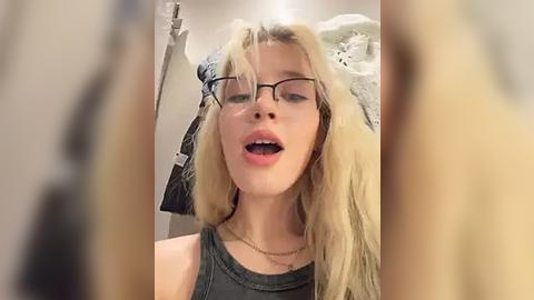 Media: Video of a blonde woman with straight hair, wearing black-rimmed glasses, a sleeveless dark top, and a gold necklace. She has an open-mouth expression, likely speaking or singing. Background shows a blurred room with clothes hanging.