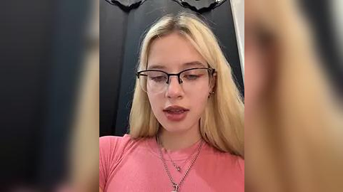 Media: Video of a young, fair-skinned, blonde woman with glasses wearing a pink top, sitting indoors against a black backdrop. She has a neutral expression.