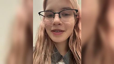 Media: A close-up video of a young woman with light skin and long, wavy pink hair, wearing black-rimmed glasses and a grey shirt.