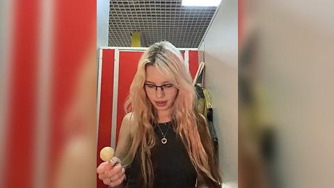 Media: Video of a woman with long, wavy blonde hair, glasses, and a black top, holding a hairbrush, standing in a narrow hallway with red walls, white ceiling tiles, and a yellow jacket hanging on a hook.