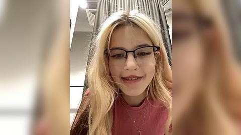 Media: Video of a smiling young girl with long blonde hair, wearing black-rimmed glasses and a pink shirt, in a brightly lit indoor setting.