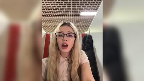 Media: Video of a young woman with long blonde hair, wearing glasses, a white blouse, and looking surprised. Background shows a tiled ceiling, red and white walls, and a black coat hanging.