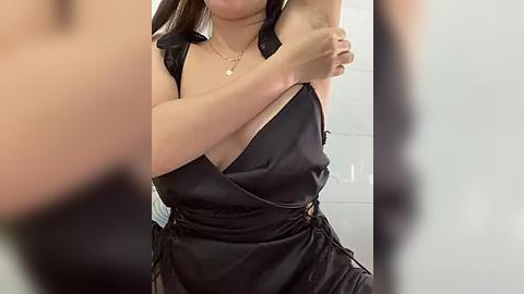 Media: A video of a woman with medium skin tone, wearing a black satin dress, adjusting her bra strap in a bathroom with white tiles.