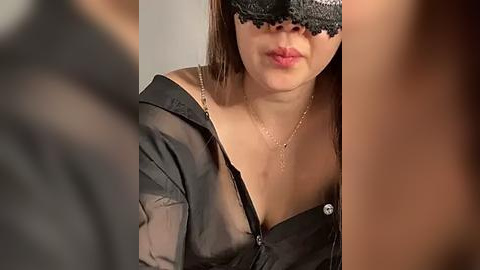 Media: A video of a woman with light skin and brown hair, wearing a black lace mask, a sheer black blouse, and a gold necklace, partially visible. The background is blurred.