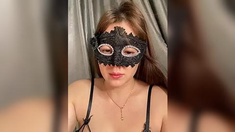 Media: Video of a young woman with long brown hair, wearing a black lace masquerade mask with intricate designs, revealing her eyes. She has a fair complexion and is wearing a delicate gold necklace. The background is a blurred, neutral-toned curtain.