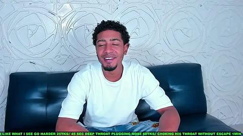 Media: Video of a muscular Black man with curly hair and a goatee, wearing a white T-shirt, sitting on a dark blue leather couch, with a textured white wall in the background. Text overlay reads: \"I lost 35lbs in 8 weeks doing the 3-second burpee workout.\