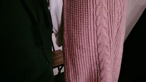 Media: Video of a person wearing a pink, textured knit sweater over a white shirt, partially visible. The sweater's intricate stitch pattern contrasts with the smooth, dark fabric of their jacket. Background includes a wooden chair and a black object.