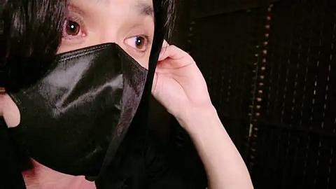 Media: A close-up video of a person with light skin and long, dark hair, wearing a black surgical mask, holding it with their right hand. The background is dimly lit, featuring a dark, textured surface.