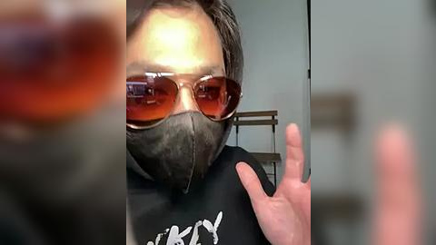 Media: Video of a man with dark sunglasses, black face mask, and black t-shirt, gesturing with his hand. Background features a blurred, metallic object.