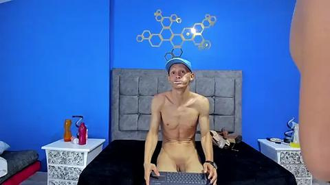 Media: Video of a naked, muscular man with a blue cap and tape over his mouth, sitting on a black bed in a blue room with science-themed wall art.