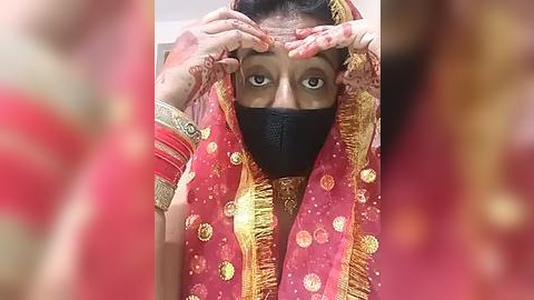 Media: A video of a woman in a red, gold-embellished sari with a black face mask, covering her nose and mouth, and applying makeup to her forehead.