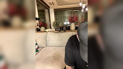 Media: Video of a modern living room with beige sectional sofa, glass-top coffee table, large windows, potted plants, and a person in a black shirt and cap.