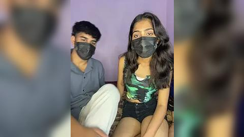 Media: Video of two young people with masks, sitting on a bed with white sheets. The woman, with long dark hair, wears a green camo crop top and black shorts. The man, with short dark hair, wears a gray shirt and white pants.