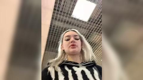 Media: Video of a blonde woman with light skin, wearing a black and white striped top, standing in a modern, industrial space with metal grids and a bright ceiling light.