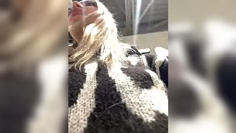 Media: A blurred video of a woman with long, wavy blonde hair wearing a black and white knit sweater, seated in a dimly lit room. Her expression is indiscernible due to the focus blur.