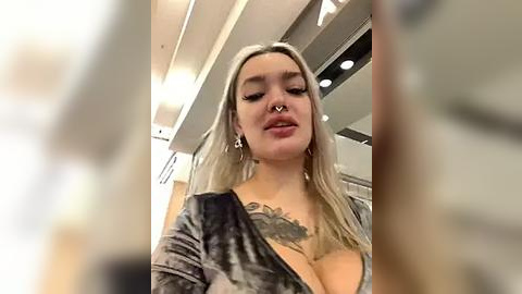 Media: Video of a young woman with long platinum blonde hair, wearing a gray velvet dress, revealing a large floral tattoo on her chest, standing in a modern, dimly lit room with recessed lighting.