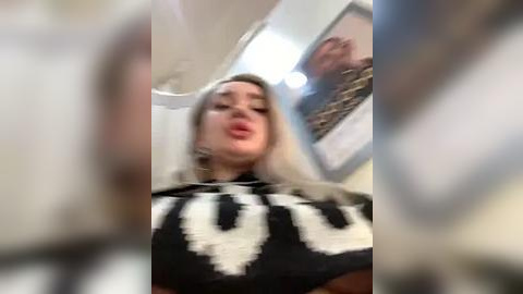 Media: A video of a woman with long, straight blonde hair, wearing a black and white patterned top, taken indoors. The image is blurry, obscuring details.