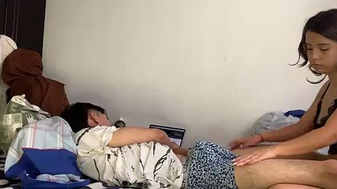 Media: Video of a young boy in a white striped shirt and blue shorts lying on a bed, while a woman in a black top and blue patterned shorts massages his leg in a simple bedroom.