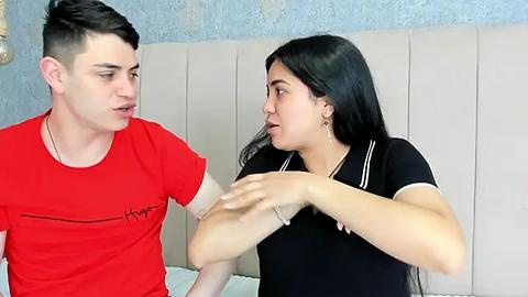 Media: Video of a young man with short dark hair in a red t-shirt and a woman with long black hair in a black blouse, both in a bedroom with a light blue wall and white headboard.