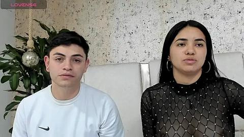 Media: Video of a young man with short dark hair and a white t-shirt, and a young woman with long black hair and a black sequined top, standing side by side in a modern room with a white headboard and a hanging plant.