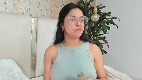 Media: Video of a Latina woman with long black hair and fair skin, wearing large eyeglasses and a light blue sleeveless top, sitting on a bed with a white headboard, holding a green bottle.