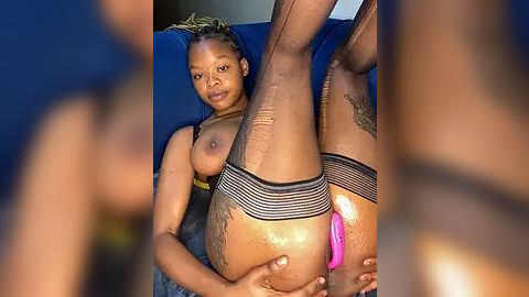 Media: Video of a Black woman with braided hair, lying on a blue couch, legs raised, wearing black fishnet stockings, exposing her vulva with a pink dildo inserted.