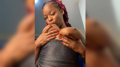 Media: Video of a Black woman with a medium skin tone and red braided hair, wearing a gray, textured dress. She is holding her bare breasts with her hands, with red-painted nails, in a dimly lit room.