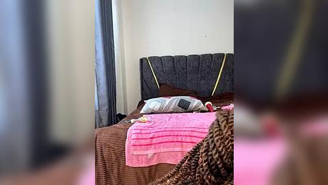 Media: Video of a simple bedroom with a brown bed, pink blanket, and a gray pillow, set against a white wall with a dark curtain to the left.