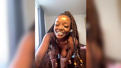 Media: Video of a joyful, topless Black woman with long, braided hair, leaning forward, smiling in a dimly lit bedroom.