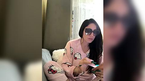 Media: Video of a young woman with long black hair, wearing sunglasses and pink pajamas with Hello Kitty print, sitting on a bed, holding a smartphone.