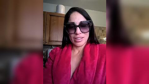 Media: A video of a smiling woman with long dark hair, wearing oversized black sunglasses and a red robe, standing in a kitchen with wooden cabinets and a beige tiled backsplash.