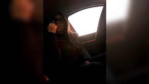 Media: Video of a woman with long hair and dark sunglasses, sitting in a dimly lit car, holding a camera, looking out the window at a bright, sunlit scene.