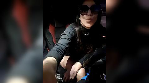 Media: Video of a woman with long dark hair, wearing sunglasses and a black top, sitting in a car with red seats.