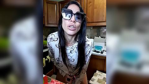 Media: Video of a woman with long black hair and oversized sunglasses in a kitchen, wearing a white top with black cat prints.