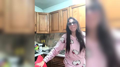 Media: Video of a smiling woman with long dark hair in a pink Hello Kitty pajama shirt, standing in a kitchen with wooden cabinets and a cluttered countertop.