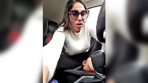 Media: Video of a woman in a car seat, wearing sunglasses and a white sweater, looking distressed, with blurred figures in the foreground.